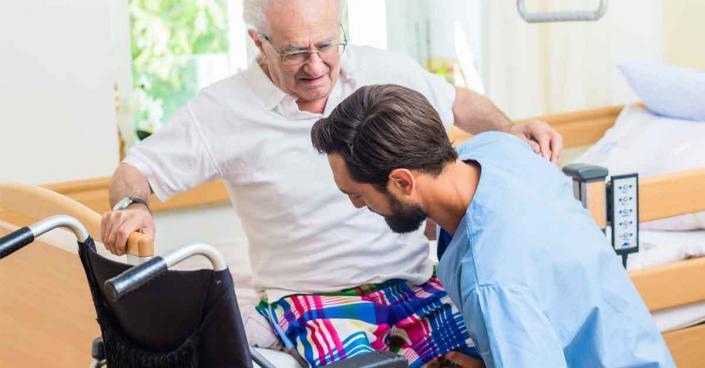 Altenpfleger hilft aelterem Mann aus Rollstuhl ins Bett